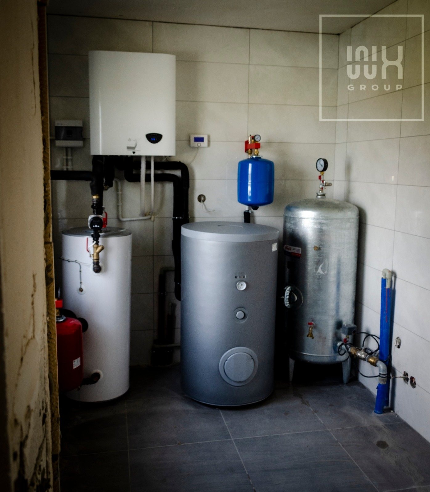 Kotłownia z zainstalowanymi zbiornikami ciepłej wody, kotłem i naczyniami wzbiorczymi w pomieszczeniu wyłożonym białymi płytkami, widoczne rury i armatura, w rogu logo firmy.