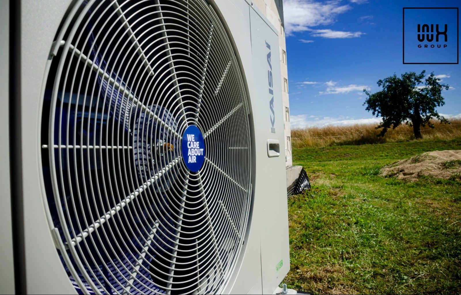Zewnętrzna jednostka klimatyzacji Kaisai z widocznym wentylatorem i naklejką 'We care about air', na tle zielonej trawy i błękitnego nieba z drzewem, logo firmy w prawym górnym rogu.