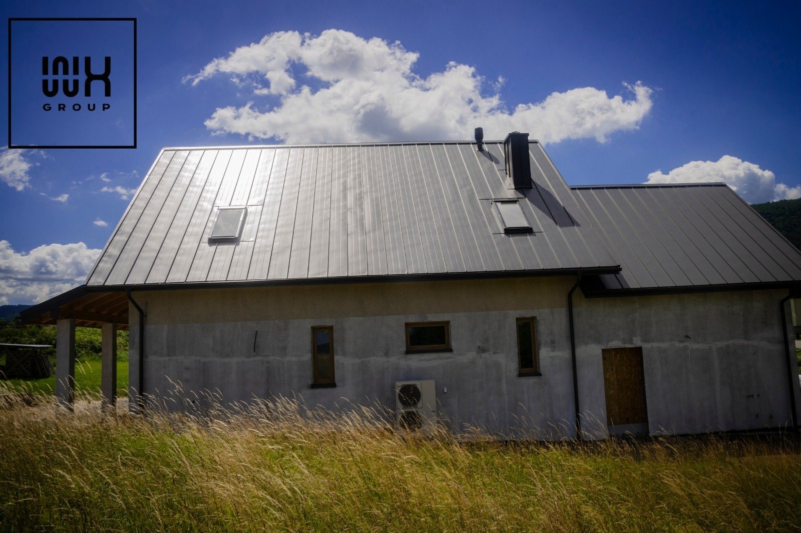 Nowoczesny dom jednorodzinny z szarą blachą na dachu, widocznym kominem, oknami dachowymi i klimatyzatorem na ścianie, otoczony wysoką trawą w słoneczny dzień.