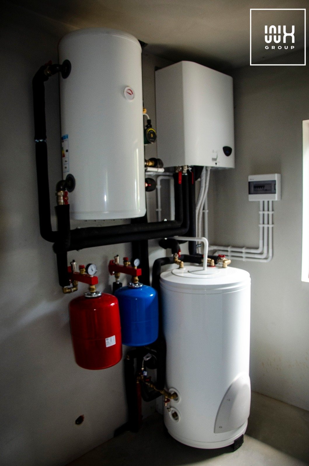 Zestaw urządzeń grzewczych: dwa białe zbiorniki, czerwony i niebieski zbiornik wyrównawczy, czarne rury, biała skrzynka sterownicza na szarej ścianie, logo firmy w prawym górnym rogu.