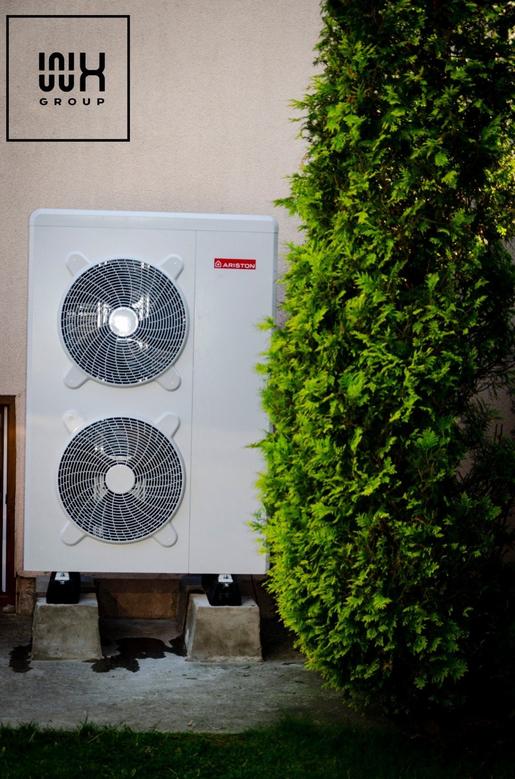 Biała pompa ciepła marki Ariston, zamontowana na betonowych podstawach przy ścianie budynku, obok rośnie wysoki, zielony krzew iglasty. W lewym górnym rogu widoczne logo firmy.