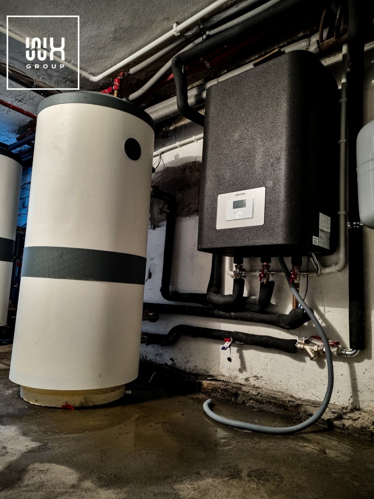 Dwa zbiorniki: biały pionowy z czarnym pasem i czarny prostopadłościenny z panelem sterowania, połączone rurami w kotłowni z wilgotną podłogą.