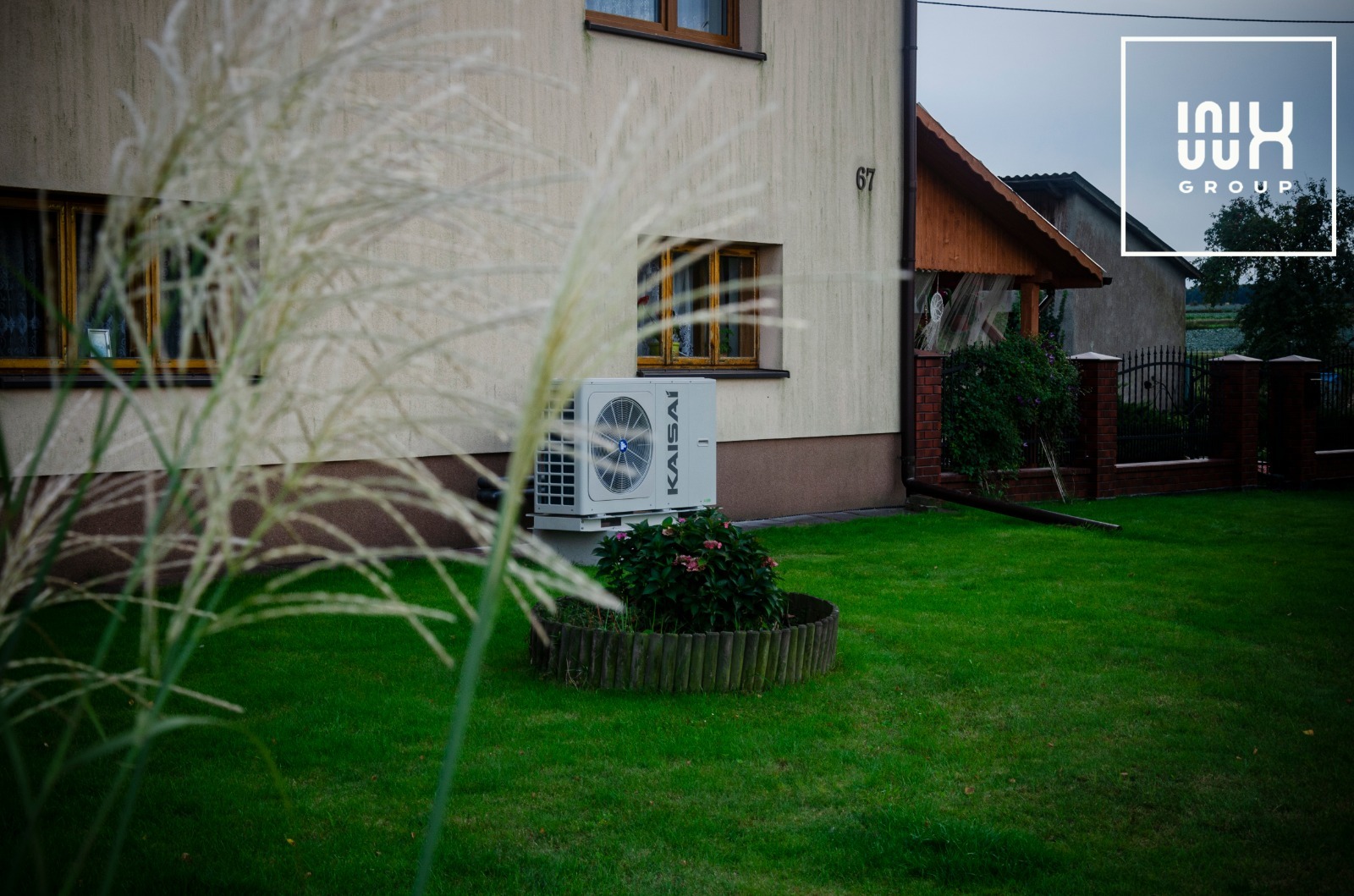 Biała jednostka zewnętrzna klimatyzacji Kaisai zamontowana na ścianie domu z numerem 67, widoczna zza ozdobnej trawy na trawniku.