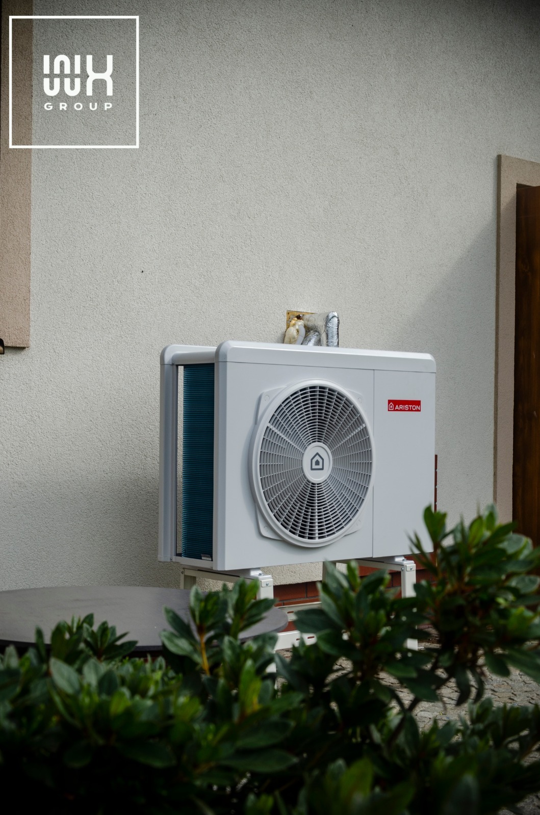 Zewnętrzna jednostka pompy ciepła Ariston zamontowana na białej ścianie budynku, widoczna zza zielonych krzewów, z fragmentem brązowych drzwi w tle.