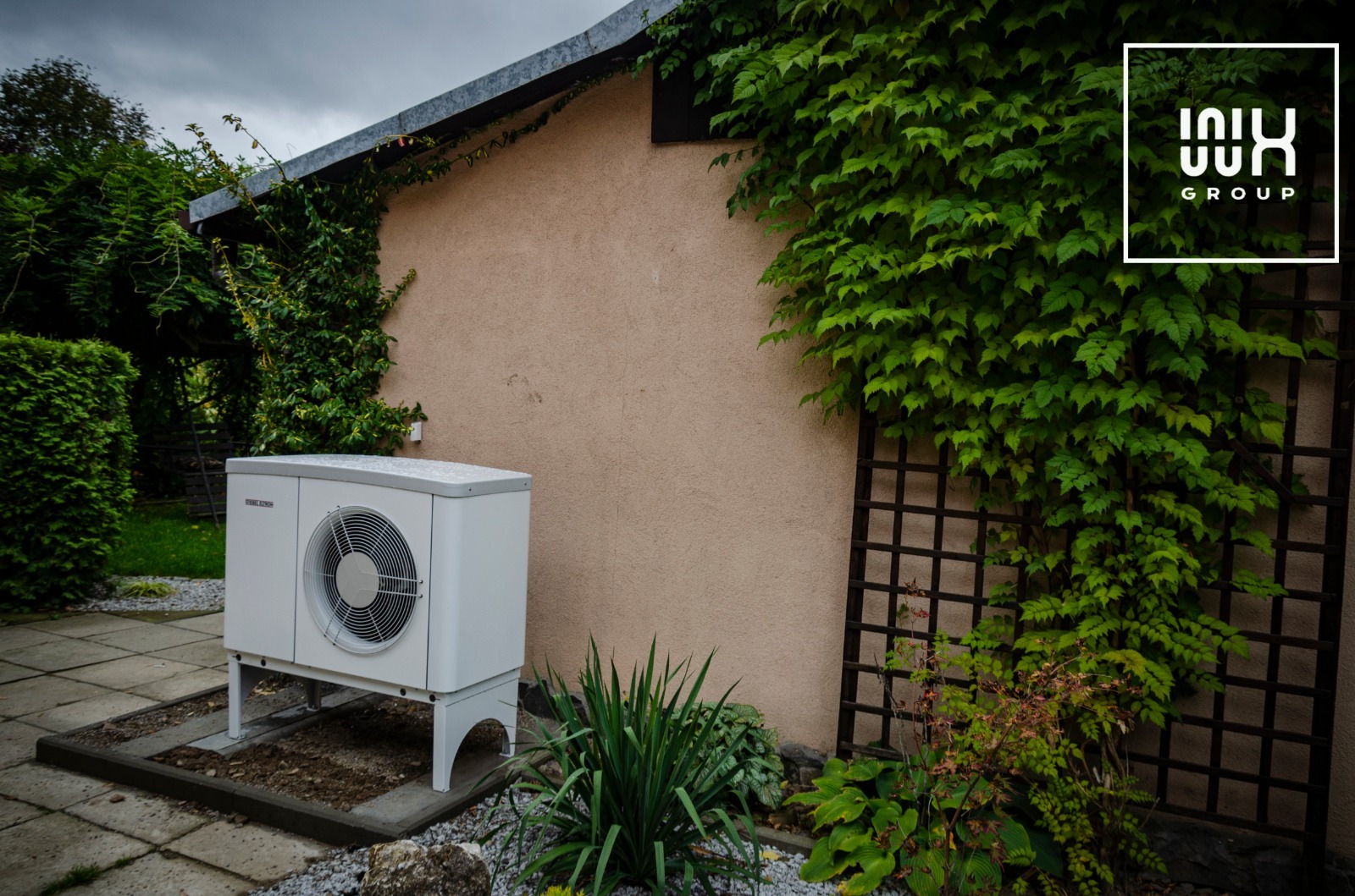 Biała pompa ciepła marki Stiebel Eltron zainstalowana na zewnątrz budynku z beżową elewacją, obok drewnianej kraty porośniętej zielonym bluszczem, logo firmy w prawym górnym rogu.