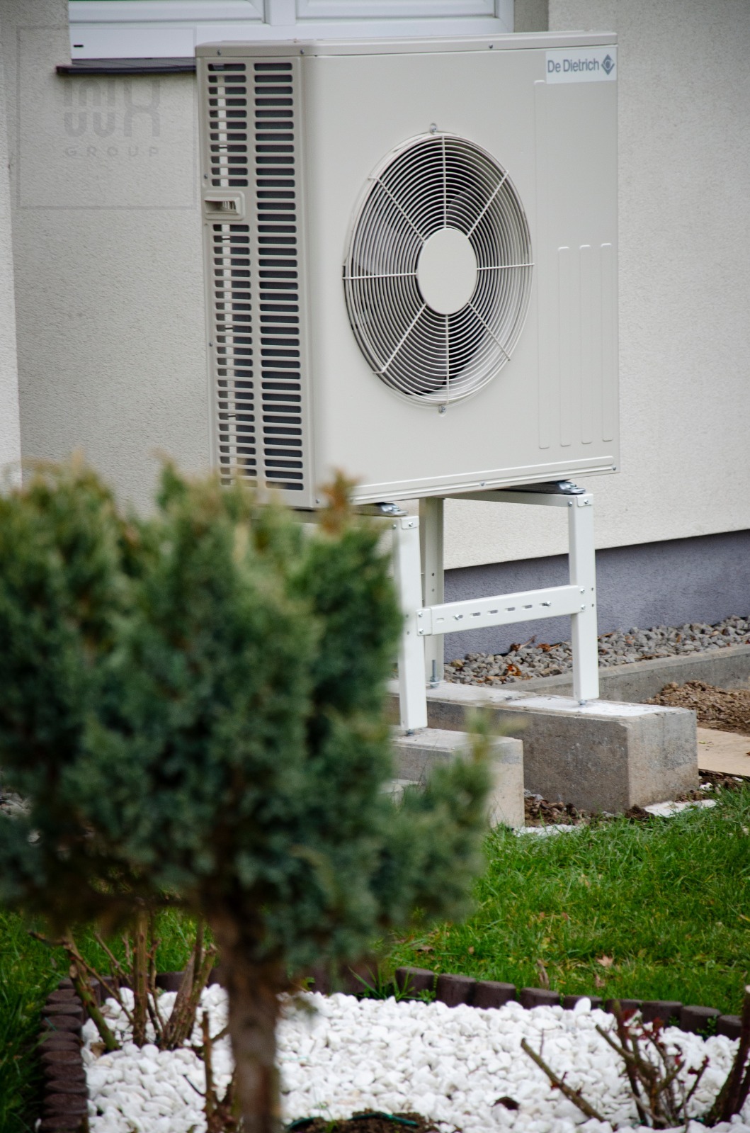 Zewnętrzna jednostka pompy ciepła De Dietrich zamontowana na białym stelażu, widoczna zza krzewu i rabaty z białymi kamykami.