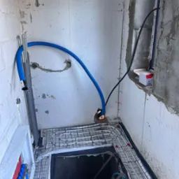 Wnętrze w trakcie instalacji ogrzewania podłogowego: białe rurki grzewcze ułożone na siatce, niebieski peszel doprowadzony do ściany, widoczna rozdzielnica z zaworami w kolorach czerwonym...