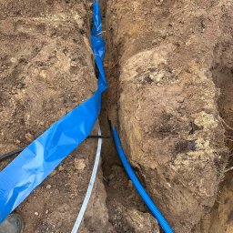 TimiElektro - Wykop w ziemi z ułożonymi przewodami elektrycznymi, zabezpieczonymi niebieską folią i niebieską rurą karbowaną, widoczna stopa buta dla skali.