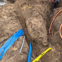 TimiElektro - Wykop z ułożonymi rurami instalacyjnymi: pomarańczowe, niebieskie karbowane oraz taśma ostrzegawcza GAZ, pracownik z łopatą w tle.