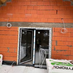 TimiElektro - Montaż okien w surowym stanie: widoczna czerwona cegła dziurawka, okna z czarnymi ramami z białymi napisami OKNOPLAST i VEKA, białe kable elektryczne zwisające ze ściany oraz styropian TYROPOL...