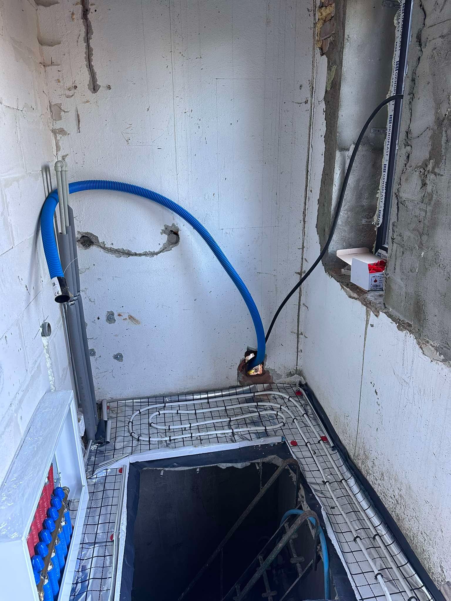 Wnętrze w trakcie instalacji ogrzewania podłogowego: białe rurki grzewcze ułożone na siatce, niebieski peszel doprowadzony do ściany, widoczna rozdzielnica z zaworami w kolorach czerwonym...