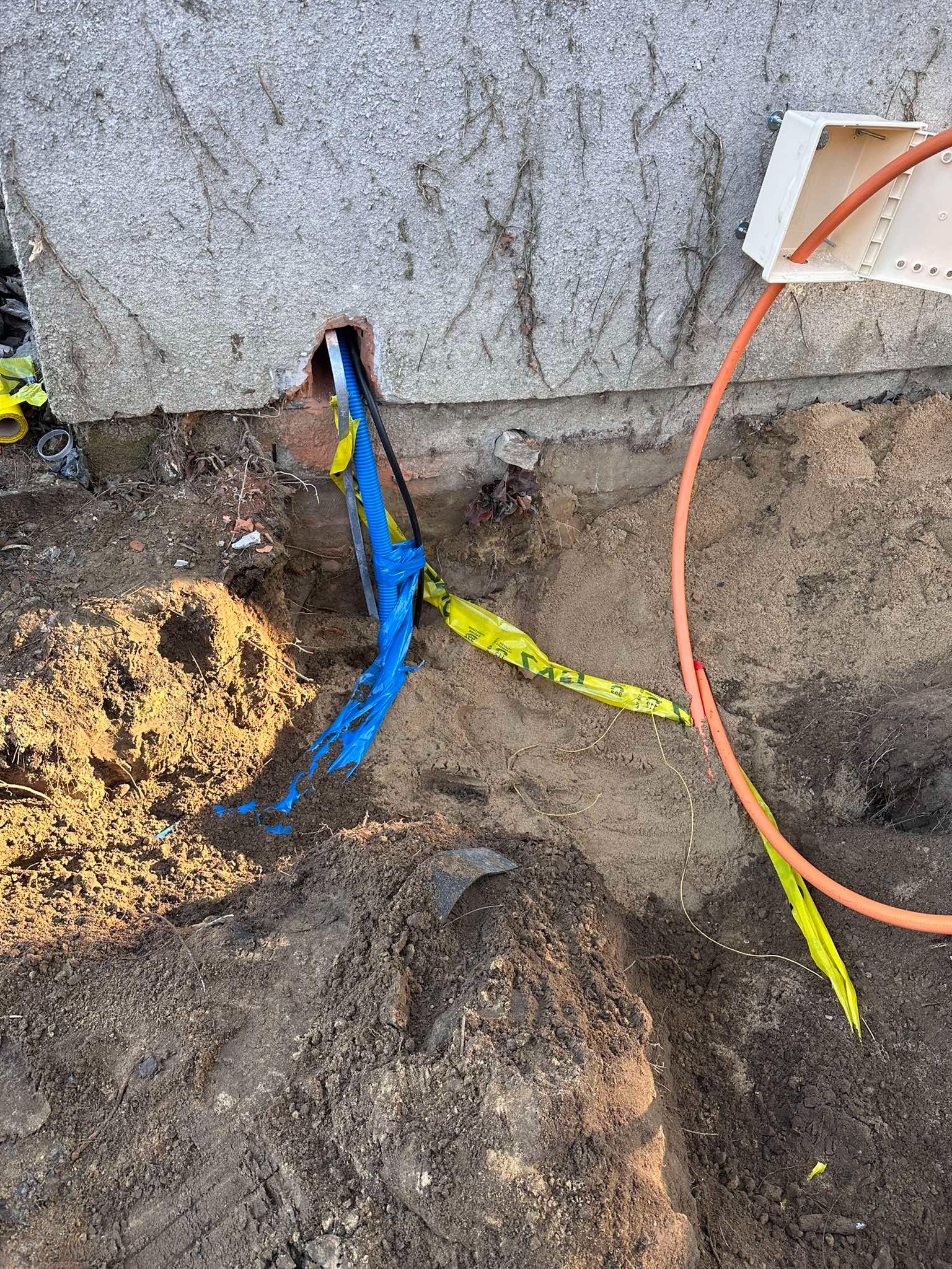 Wykop ziemny przy fundamencie budynku z wystającymi przewodami w niebieskiej karbowanej rurze i pomarańczowym kablu, zabezpieczonymi żółtą taśmą ostrzegawczą. Widoczna skrzynka instalacyjna...