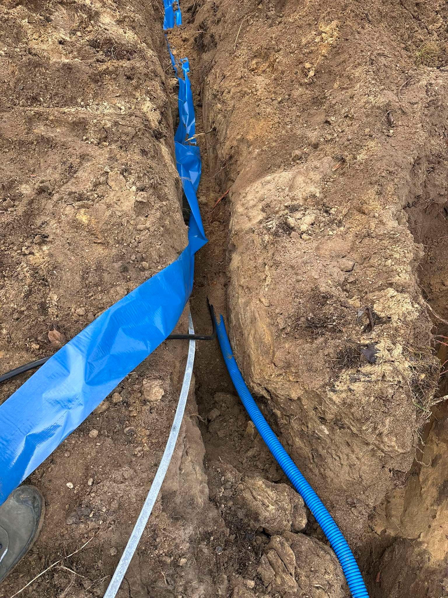 Wykop w ziemi z ułożonymi przewodami elektrycznymi, zabezpieczonymi niebieską folią i niebieską rurą karbowaną, widoczna stopa buta dla skali.