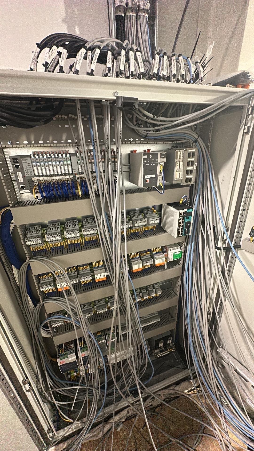 Rozdzielnica elektryczna w trakcie montażu, widoczne liczne przewody, moduły sterujące i złączki, część przewodów oznaczona etykietami, widok z góry na wnętrze szafy sterowniczej.