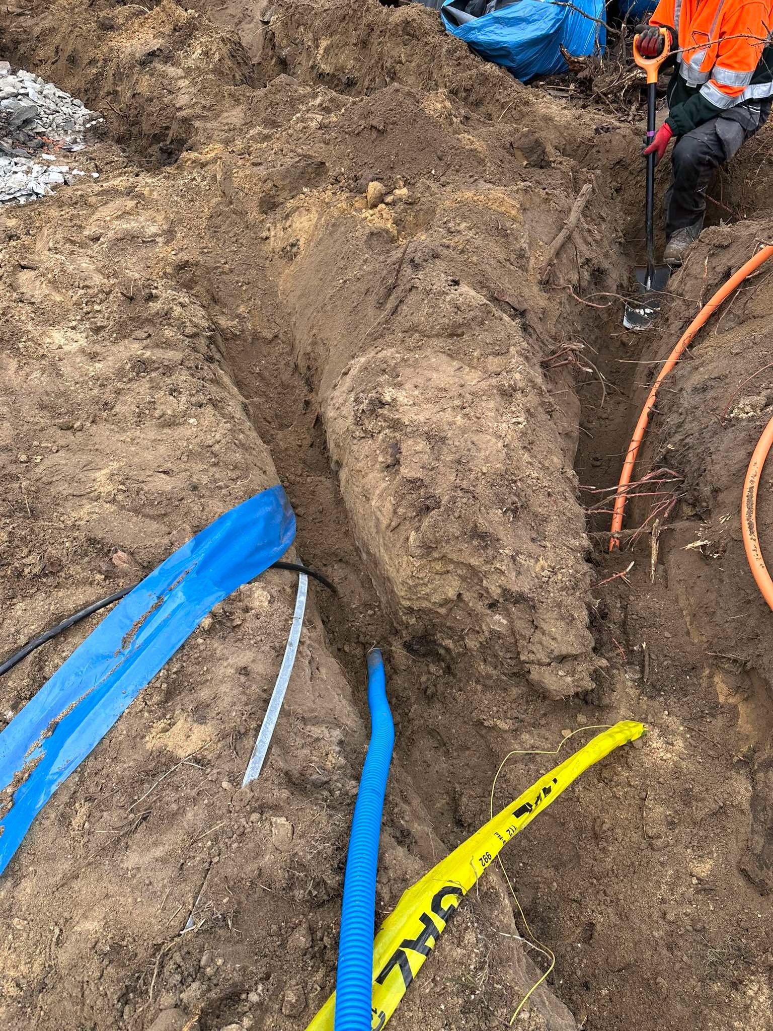 Wykop z ułożonymi rurami instalacyjnymi: pomarańczowe, niebieskie karbowane oraz taśma ostrzegawcza GAZ, pracownik z łopatą w tle.