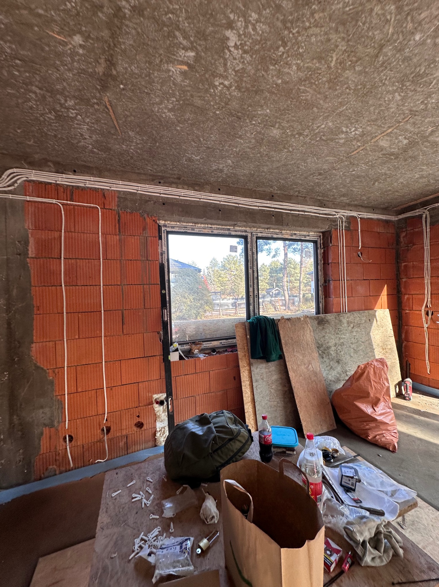 Surowy stan wnętrza w trakcie prac: widoczne przewody elektryczne prowadzone po ścianach z czerwonej cegły, okno z widokiem na zewnątrz, oraz prowizoryczny stół z narzędziami i materiałami...