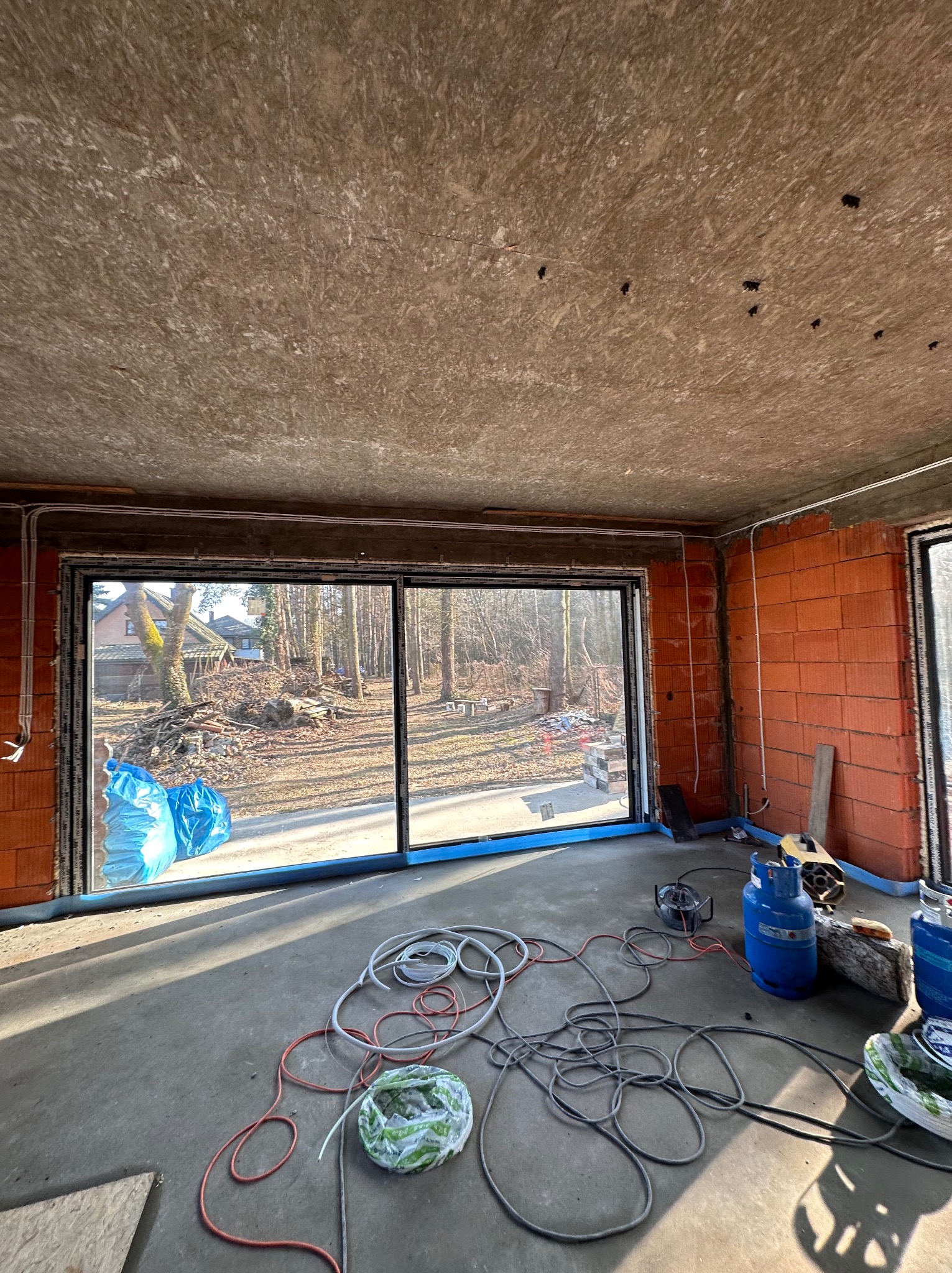 Surowy stan wnętrza z widocznymi przewodami elektrycznymi na ścianach, niezabezpieczonymi otworami w suficie, dużym oknem tarasowym i butlami gazowymi na podłodze.