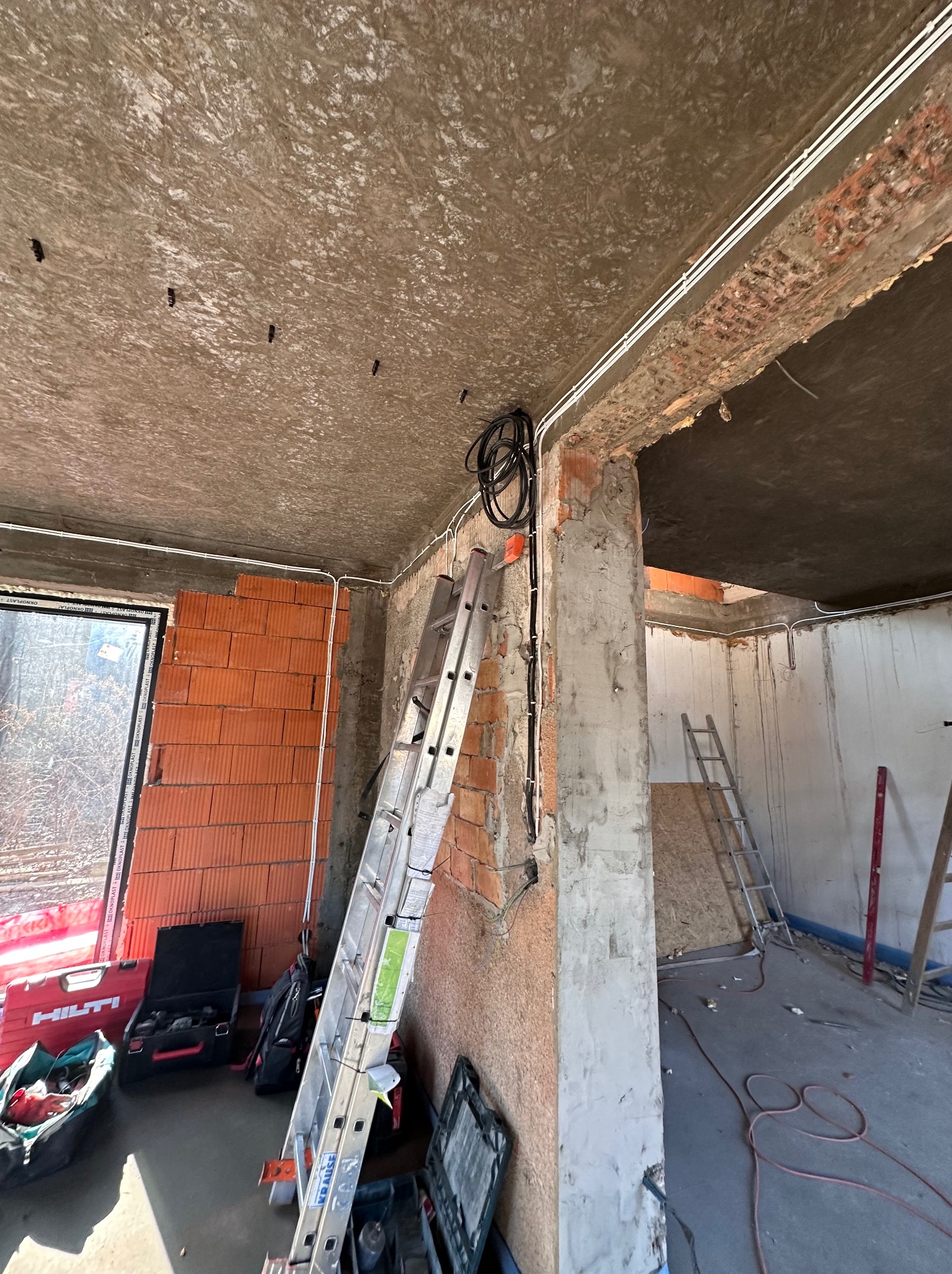 Wnętrze w trakcie remontu z widocznymi przewodami elektrycznymi prowadzonymi po ścianach i suficie, drabina oparta o ścianę, narzędzia w skrzynkach na podłodze.