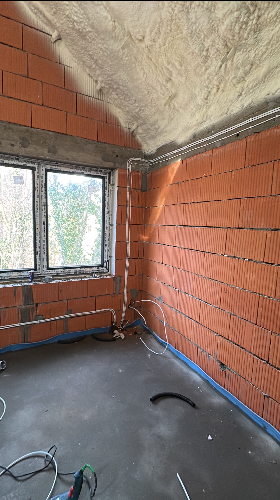 Surowy stan wnętrza z widocznymi przewodami elektrycznymi na ceglanej ścianie i izolacją piankową na suficie, okno z widokiem na drzewa, narzędzia na szarej podłodze.