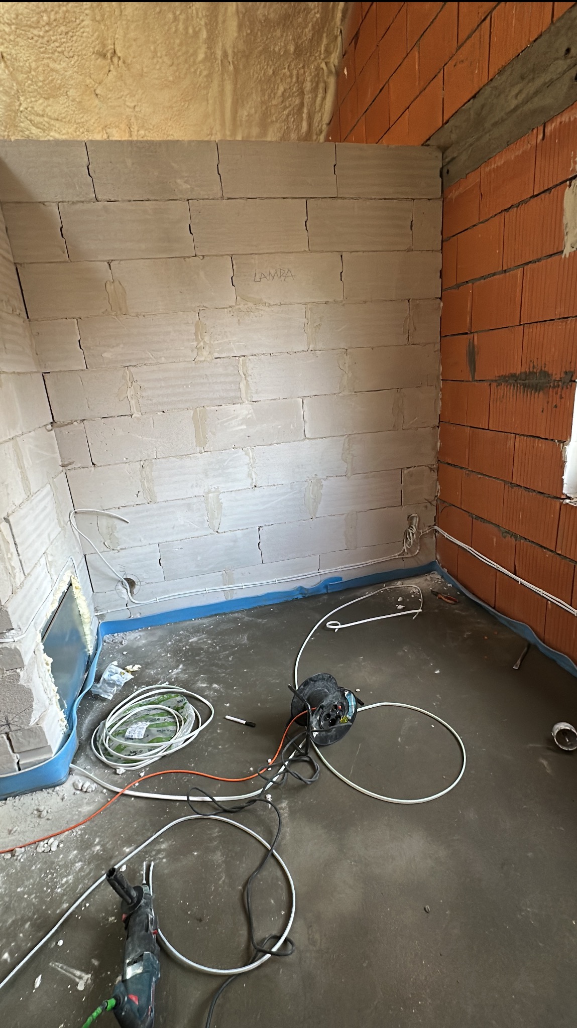 Wnętrze w trakcie prac instalacyjnych: widoczne kable elektryczne ułożone wzdłuż ścian z betonu komórkowego i cegły, narzędzia na podłodze i napis 'LAMPA' na ścianie z betonu komórkowego.