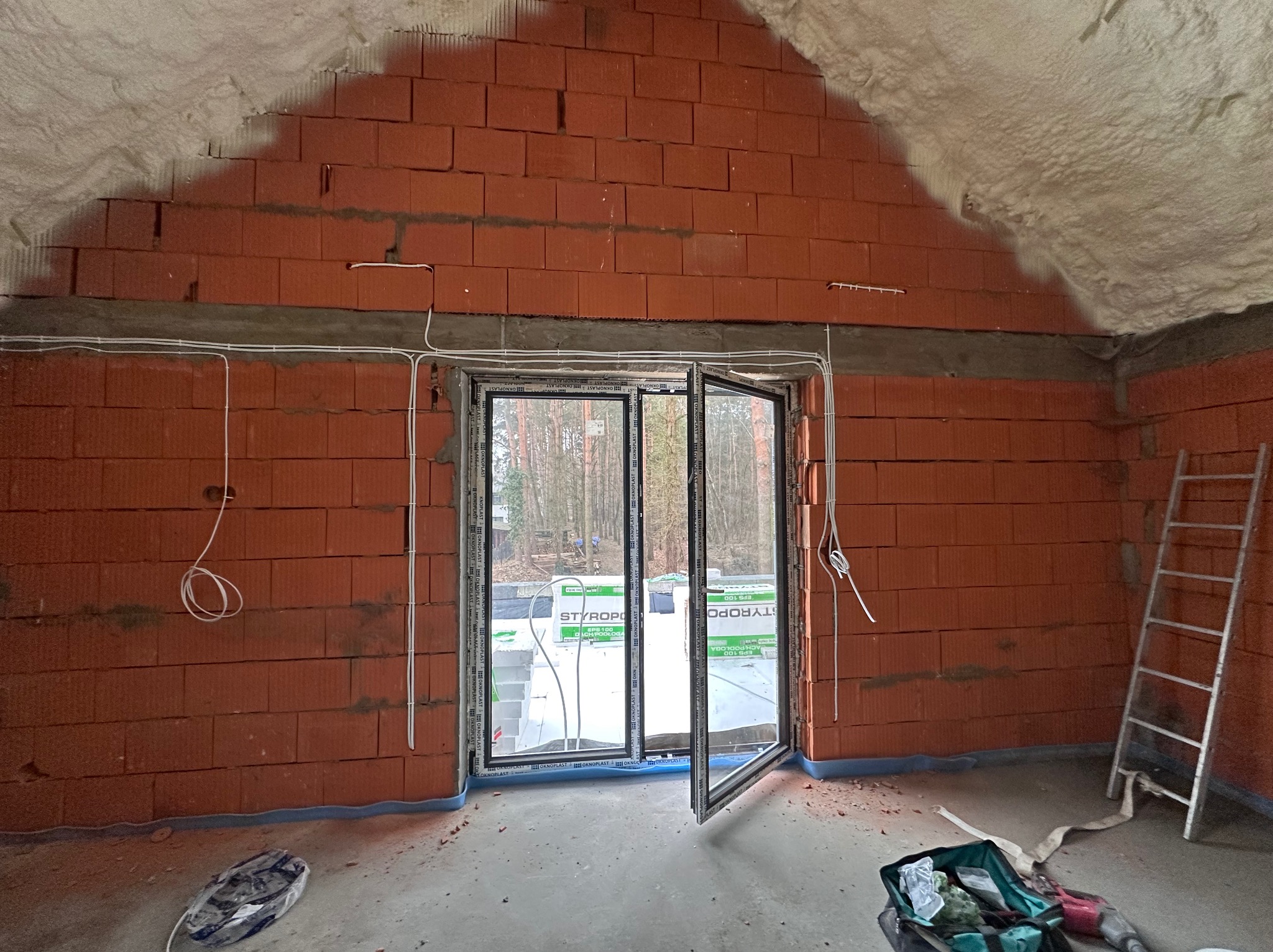 Surowy stan poddasza z oknem balkonowym, widoczne przewody elektryczne na ścianie z czerwonej cegły i ocieplenie pianą natryskową na skosach dachu.