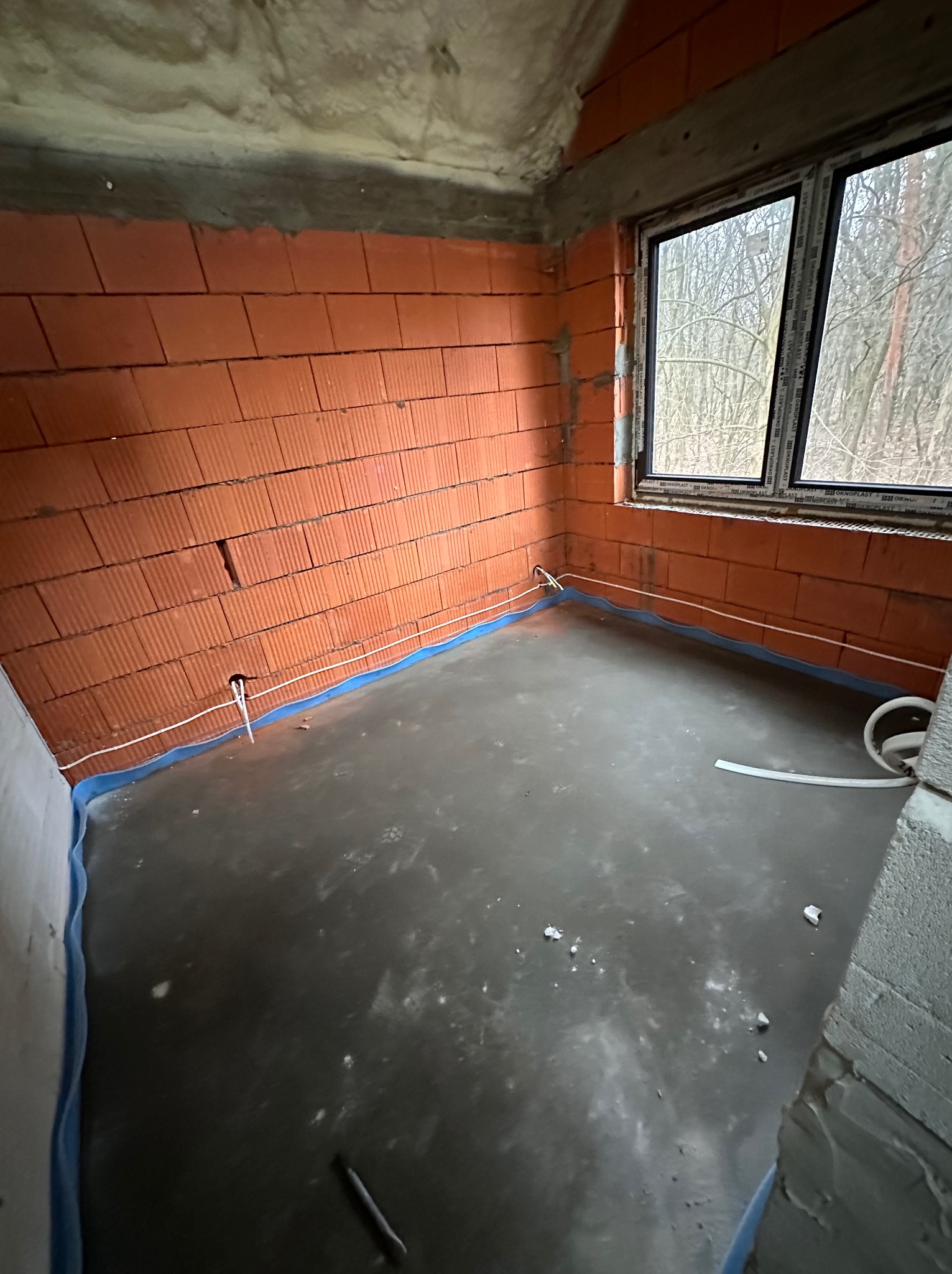 Surowy stan wnętrza pomieszczenia z ceglanymi ścianami, wylewką na podłodze, zabezpieczoną folią, i wystającymi przewodami elektrycznymi ze ściany, widok na okno z drzewami w tle.
