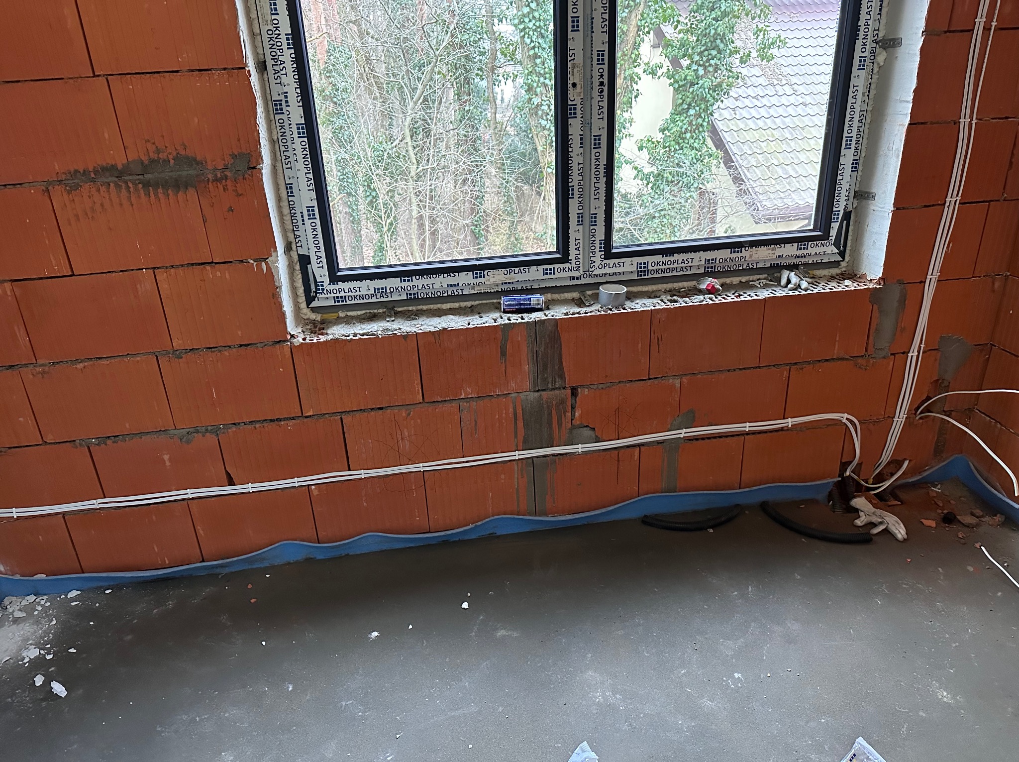 Surowy mur z czerwonej cegły ceramicznej z oknem PCV, widoczne przewody elektryczne w białej izolacji, niebieska izolacja pozioma przy podłodze, betonowa wylewka podłogowa w trakcie prac...