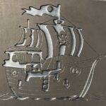 Metalowa płaskorzeźba przedstawiająca stylizowany żaglowiec z charakterystycznym, lekko groteskowym wyrazem twarzy na burcie, umieszczona na jednolitym, matowym tle.