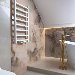DroBud Usługi Wykończeniowe Damian Drosiński - Wykończona łazienka na poddaszu z marmurowymi płytkami, złotą armaturą przy wannie wolnostojącej i białym grzejnikiem drabinkowym.