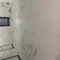 Wykończona łazienka z białymi płytkami imitującymi marmur z szarymi żyłami, czarną ramą dekoracyjną na podłodze i czarnym oknem wentylacyjnym.