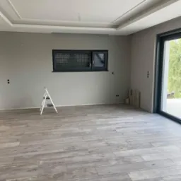 Jasne panele podłogowe ułożone w przestronnym, szarym pokoju z małym oknem i drabiną, widok na zewnątrz przez przesuwne okno balkonowe.