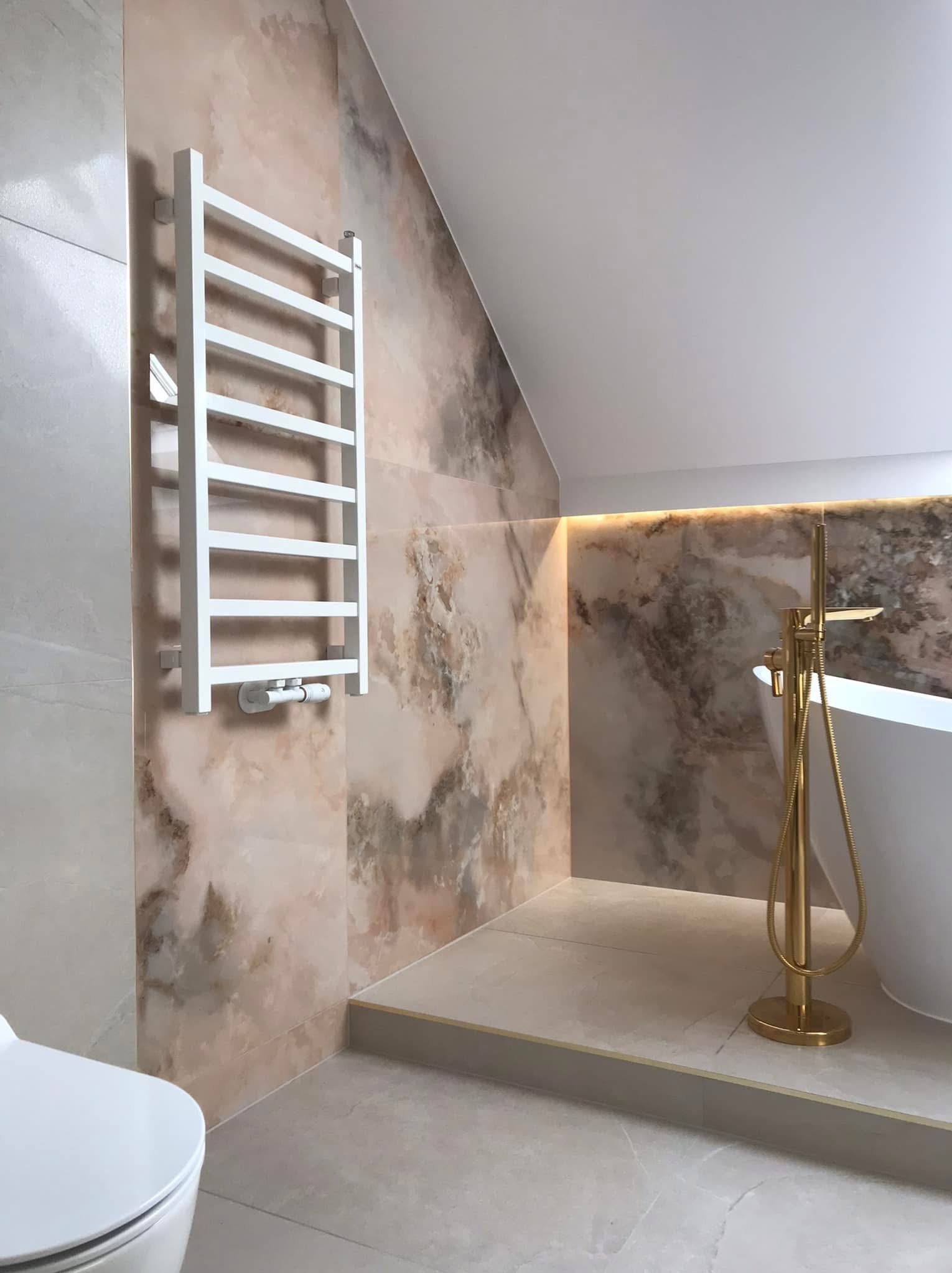 Wykończona łazienka na poddaszu z marmurowymi płytkami, złotą armaturą przy wannie wolnostojącej i białym grzejnikiem drabinkowym.
