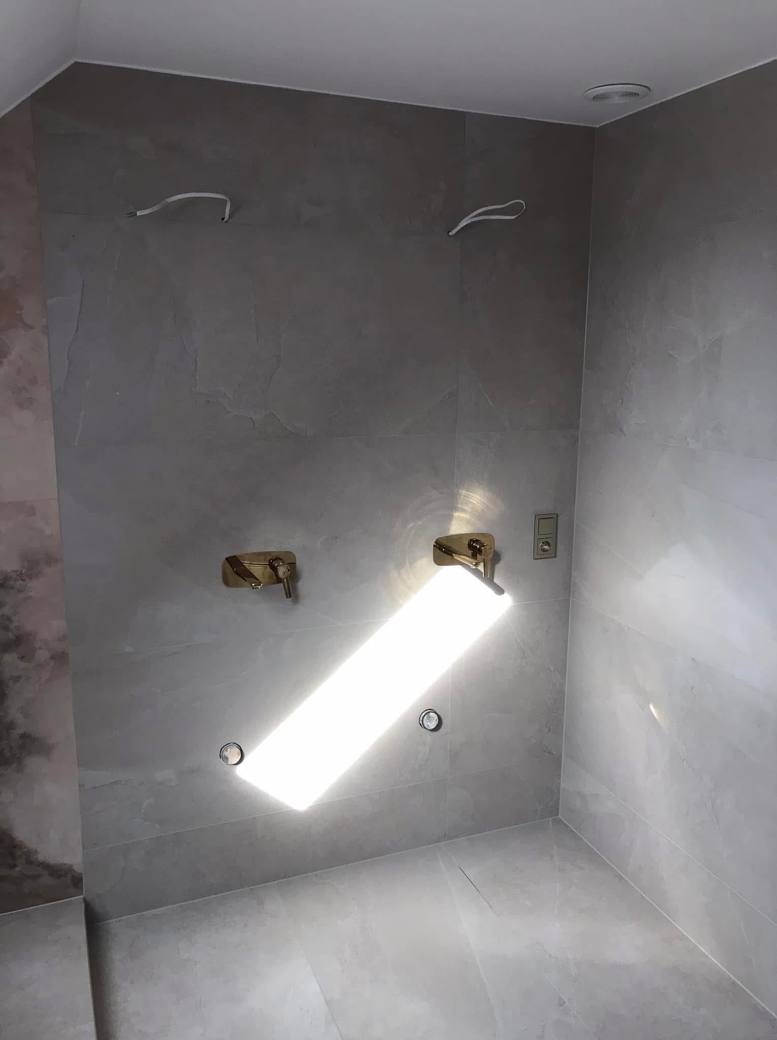 Wnętrze łazienki w trakcie remontu, wyłożonej szarymi płytkami imitującymi kamień, z zamontowaną złotą armaturą i wystającymi przewodami elektrycznymi.