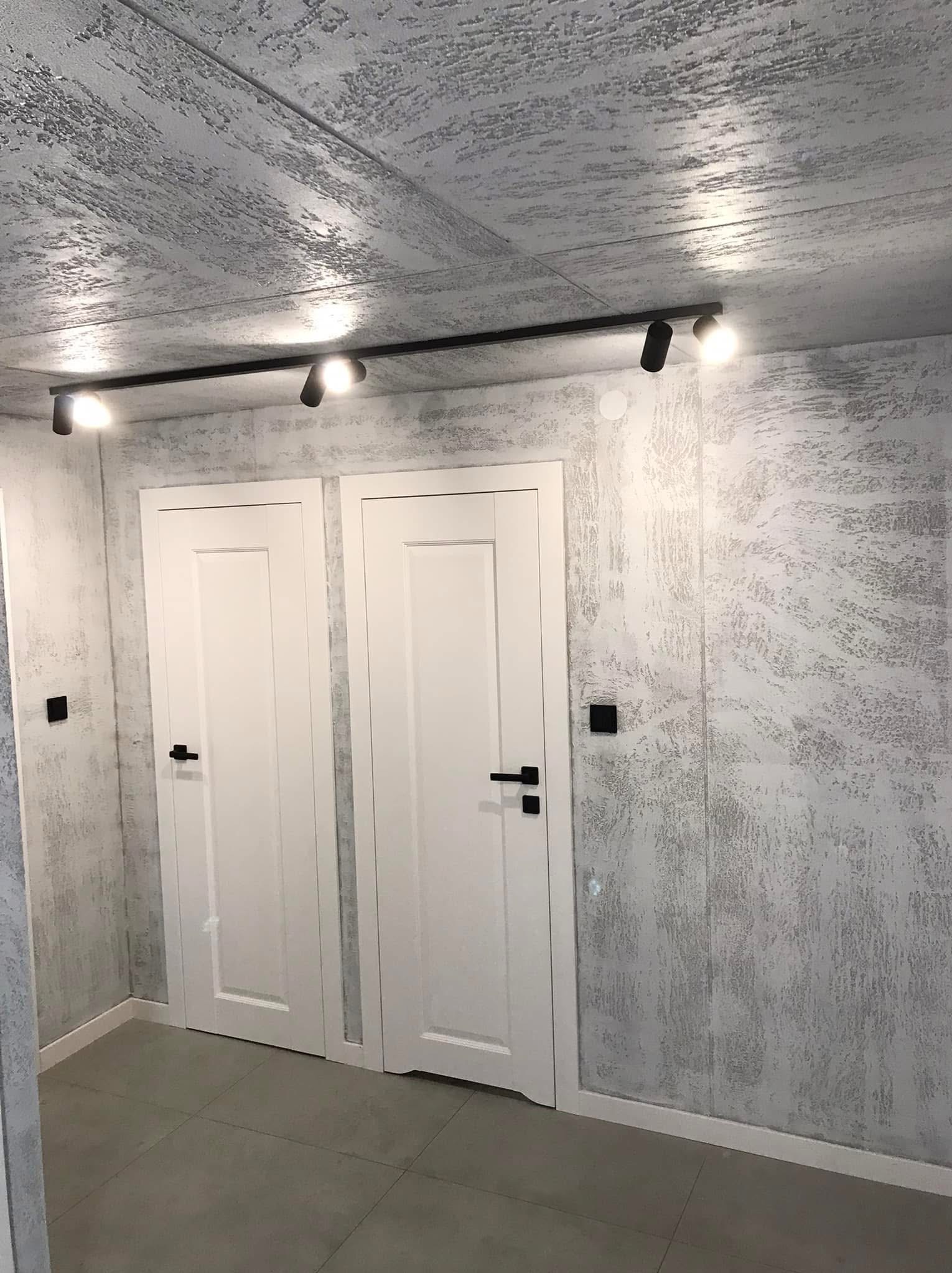 Korytarz po remoncie z dwiema białymi drzwiami, szarą podłogą z płytek i ścianami pokrytymi dekoracyjną, strukturalną farbą w odcieniach szarości, oświetlony czarną listwą z reflektorami.