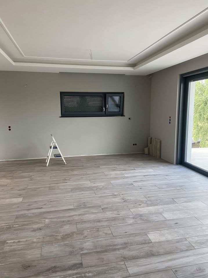 Jasne panele podłogowe ułożone w przestronnym, szarym pokoju z małym oknem i drabiną, widok na zewnątrz przez przesuwne okno balkonowe.