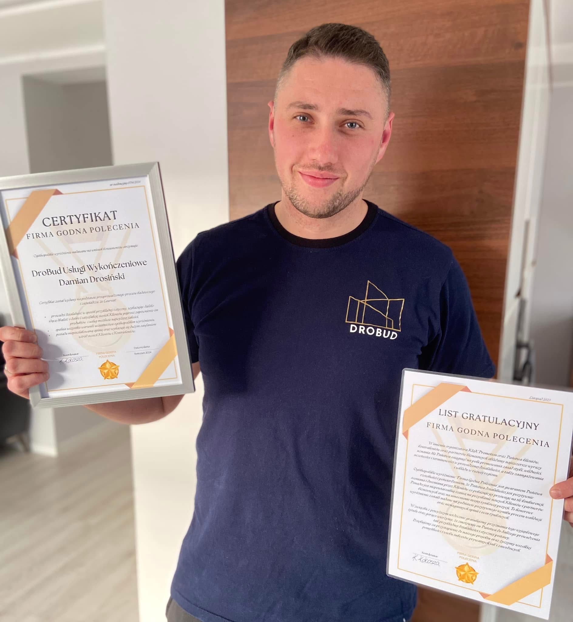 Mężczyzna w granatowej koszulce z logo firmy budowlanej DroBud prezentuje dwa certyfikaty 'Firma Godna Polecenia' i 'List Gratulacyjny', uśmiechając się do kamery.