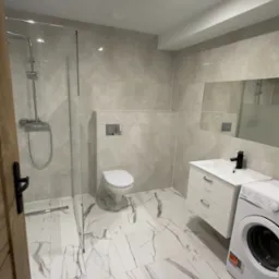 Nowoczesna łazienka z kabiną prysznicową typu walk-in, toaletą podwieszaną, umywalką z szafką i pralką, wykończona płytkami imitującymi marmur.