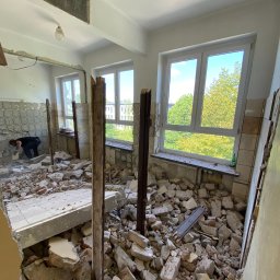 MjHorizonLine - Wnętrze w trakcie demontażu: gruz, fragmenty płytek i cegieł na podłodze, pracownik w tle skuwa resztki płytek ze ściany, widok na zieleń za oknem.