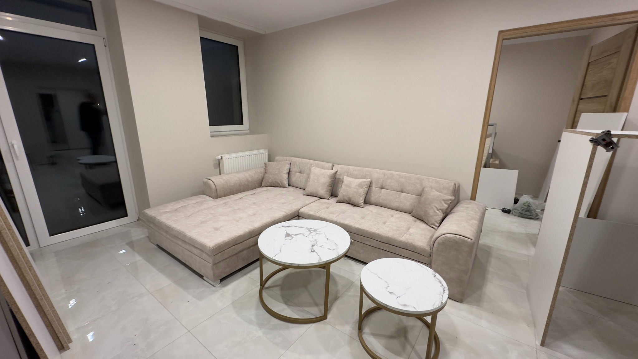 Jasny narożnik z beżową tapicerką i marmurowymi stolikami kawowymi w nowocześnie urządzonym pokoju z białymi płytkami podłogowymi, widoczne elementy zabudowy w tle.