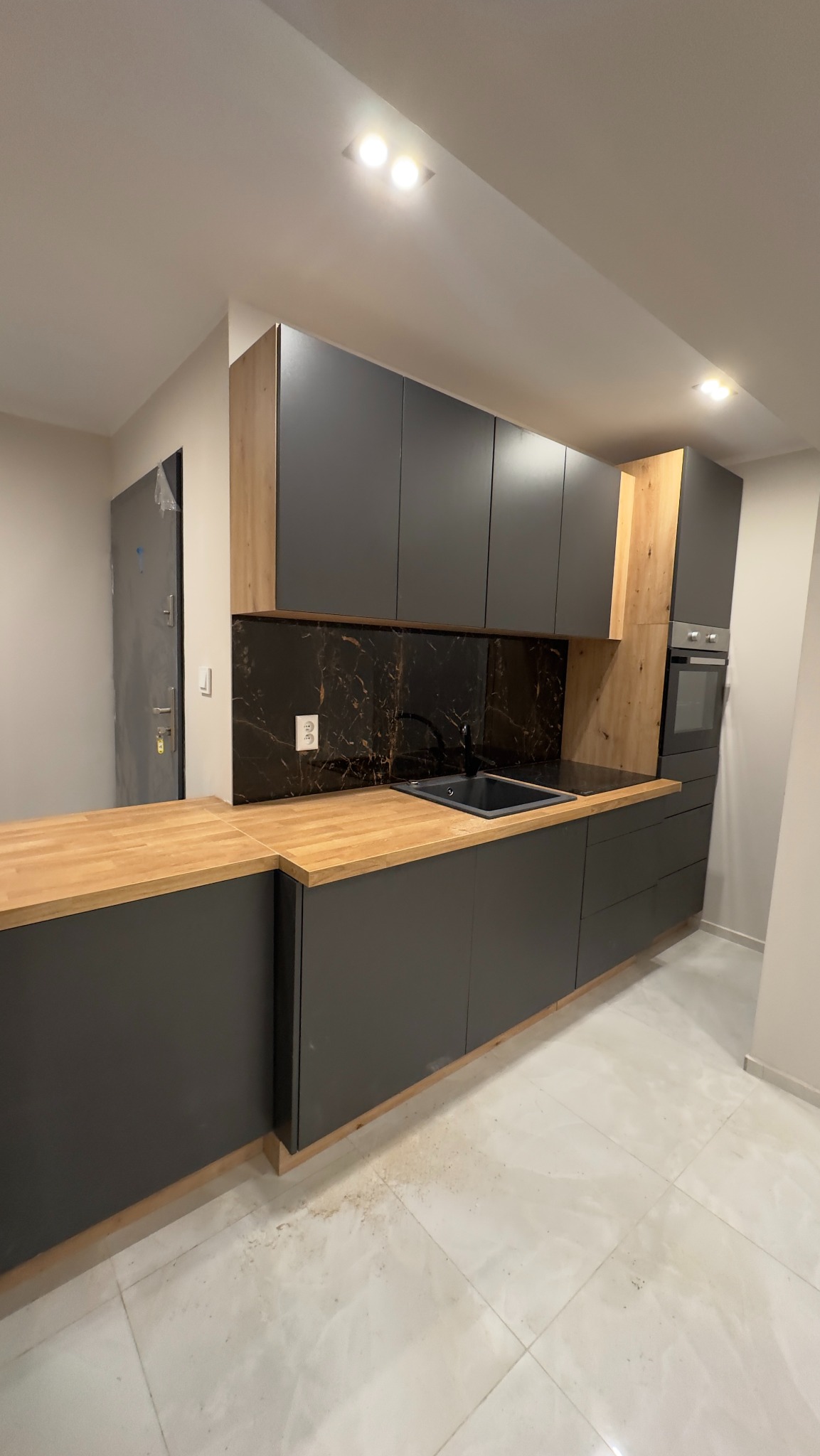 Nowoczesna kuchnia z szarymi szafkami, drewnianymi akcentami i czarnym marmurowym backsplash, widoczne gniazdko elektryczne i lekki bałagan na podłodze.