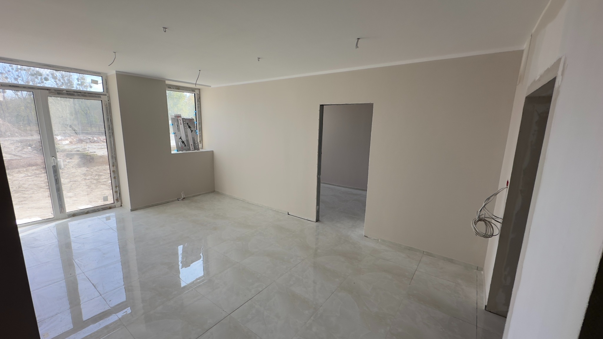 Pusty pokój po remoncie z połyskującą, marmurkową podłogą, świeżo pomalowanymi ścianami w odcieniach beżu, widocznymi przewodami elektrycznymi i oknami z widokiem na plac budowy.