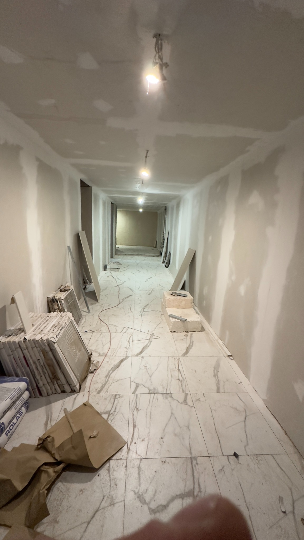 Korytarz w trakcie wykańczania: położona marmurowa podłoga, ściany przygotowane pod malowanie, widoczne worki z zaprawą i płytki ceramiczne.
