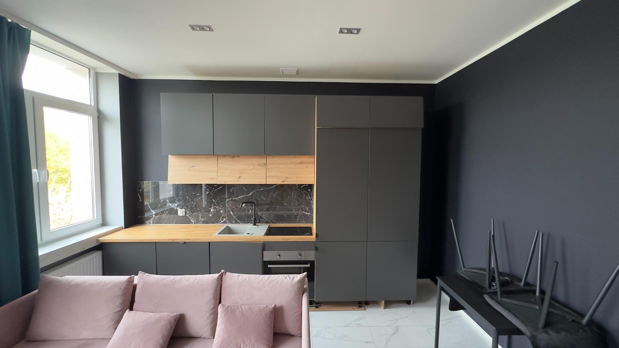 Nowoczesna kuchnia w odcieniach szarości z drewnianymi akcentami, marmurowym backsplashiem i różową sofą w pierwszym planie.