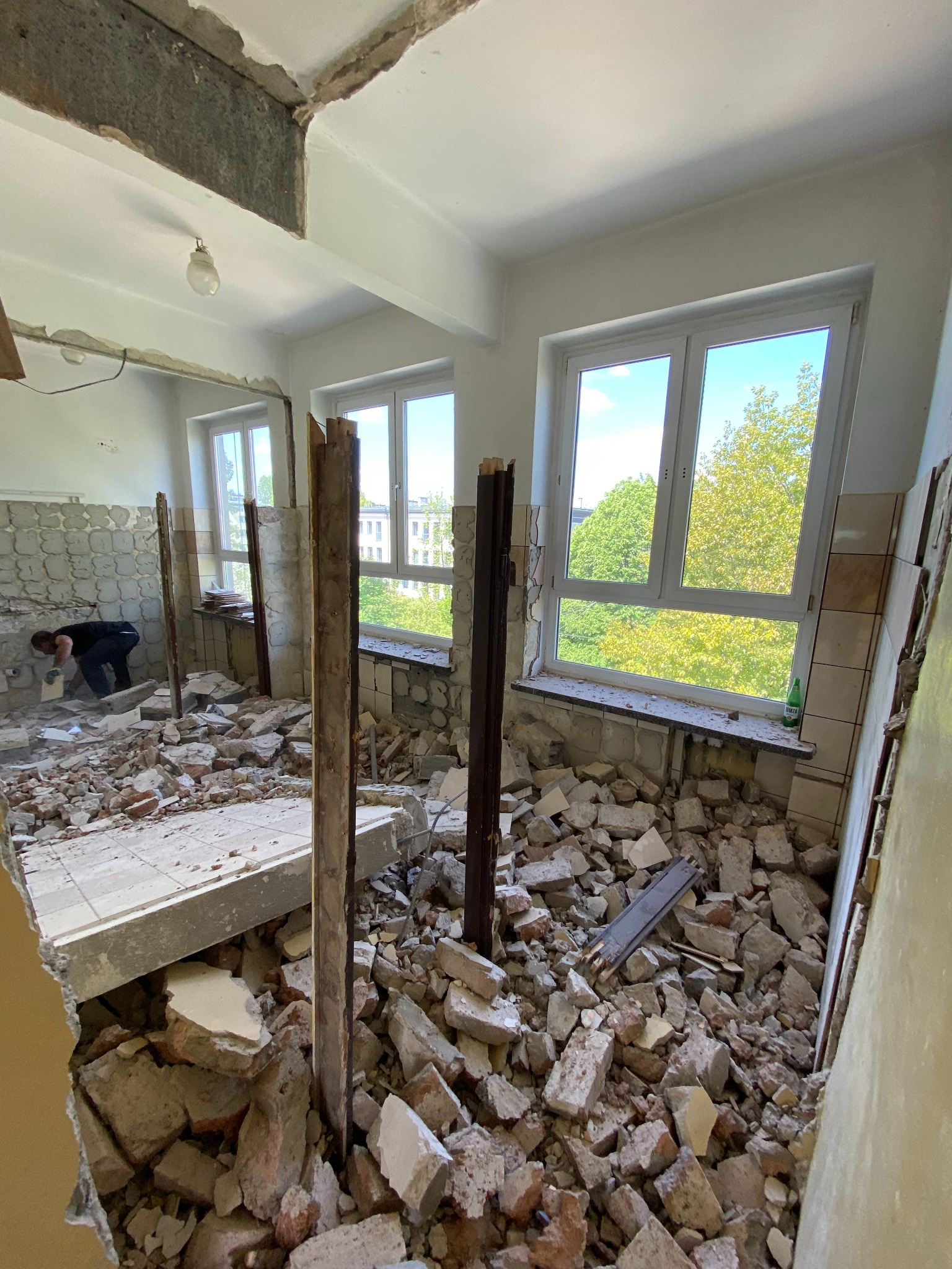 Wnętrze w trakcie demontażu: gruz, fragmenty płytek i cegieł na podłodze, pracownik w tle skuwa resztki płytek ze ściany, widok na zieleń za oknem.