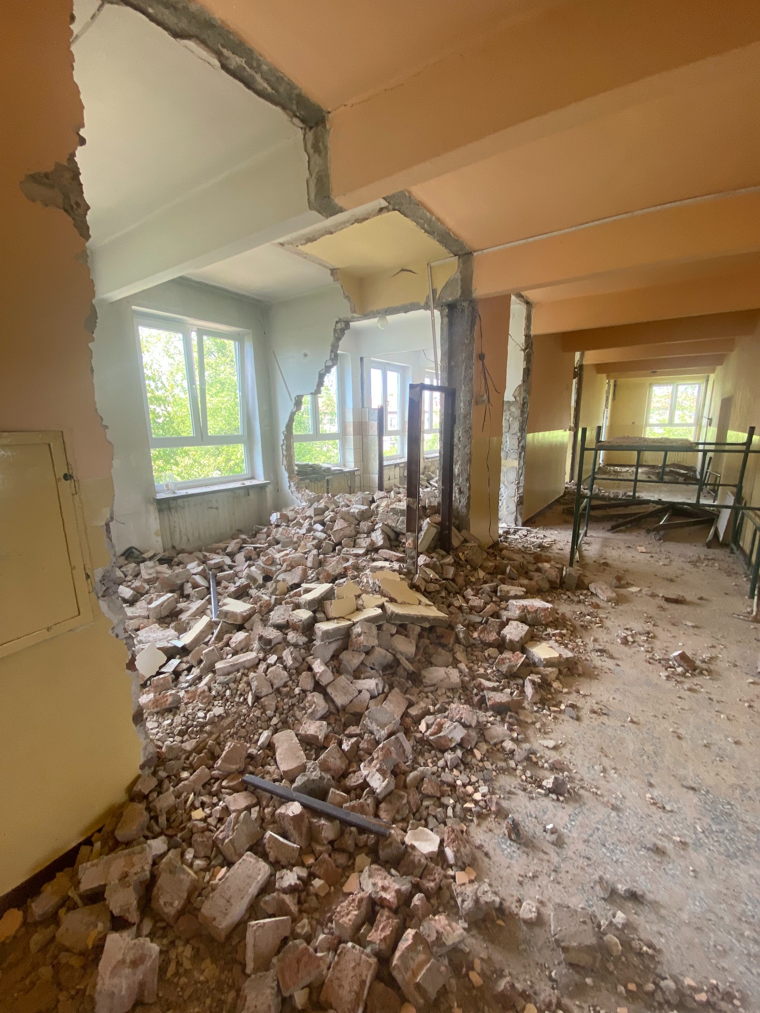 Wnętrze budynku w trakcie rozbiórki: gruz ceglany na podłodze, uszkodzone ściany i sufit, widoczne okna z zielenią na zewnątrz.