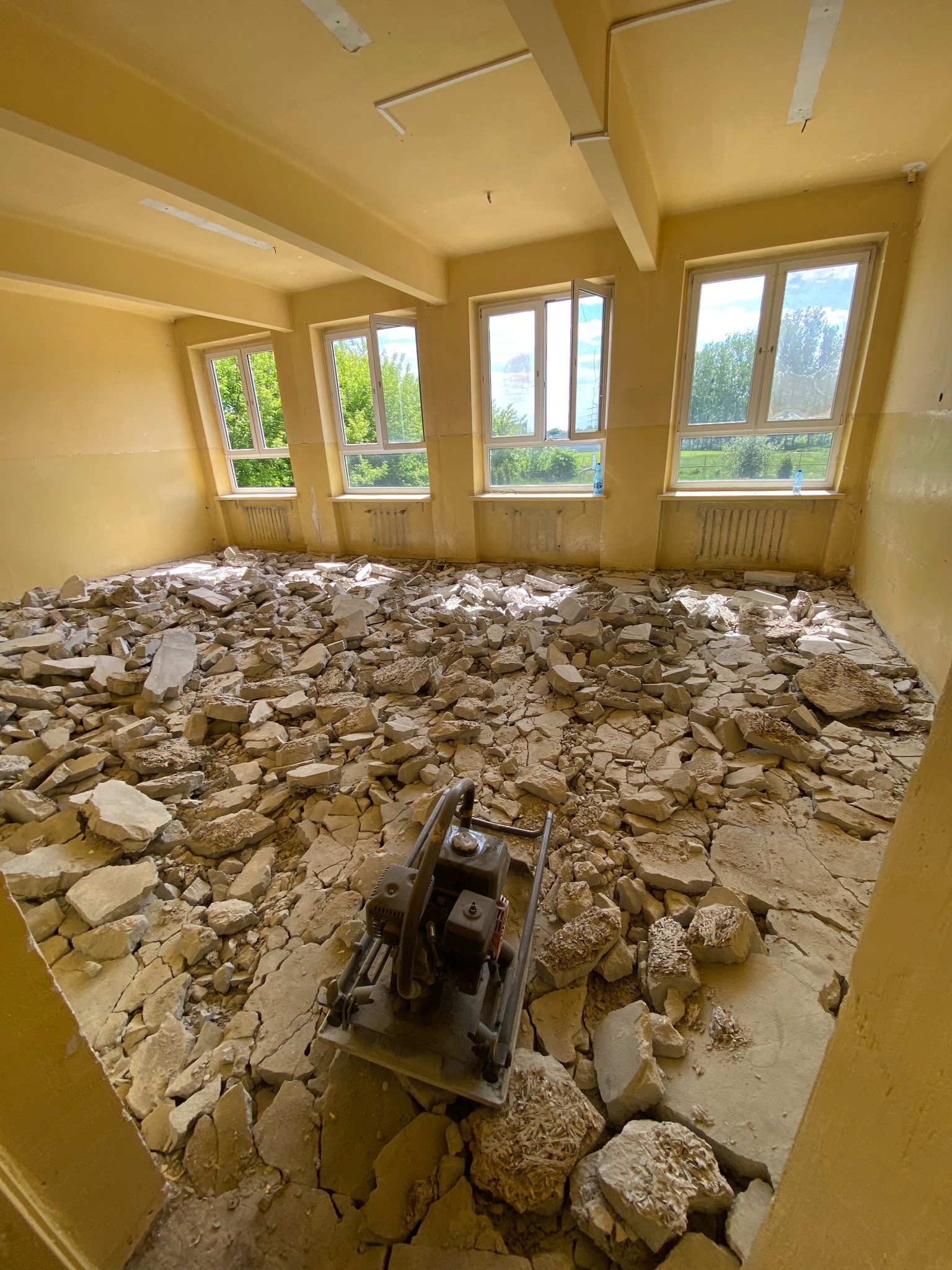 Pokój w trakcie demontażu podłogi, widoczne gruzowisko i zagęszczarka wibracyjna, żółte ściany i trzy okna z widokiem na zieleń.