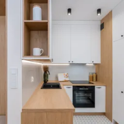 Nowoczesna kuchnia z białymi szafkami, drewnianym blatem i czarną armaturą, oświetlona punktowymi lampami sufitowymi, z widoczną książką kucharską i dekoracyjnymi słoikami.