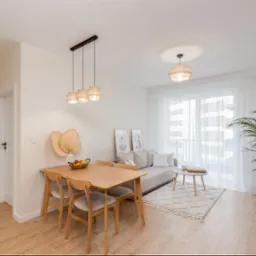 Jasne, nowocześnie urządzone wnętrze mieszkania z drewnianym stołem, krzesłami, sofą i regałem, oświetlone wiszącymi lampami z abażurami z naturalnego materiału.