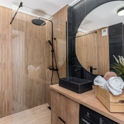 Kreator Designu - Nowoczesna łazienka z drewnopodobnymi płytkami ściennymi, czarną armaturą, okrągłym lustrem i drewnianym blatem z czarną umywalką oraz dekoracyjnym pudełkiem z ręcznikami.