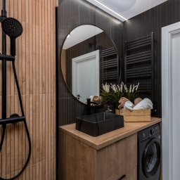 Kreator Designu - Nowoczesna łazienka z drewnopodobnymi płytkami, czarną armaturą, okrągłym lustrem i pralką pod blatem umywalki.